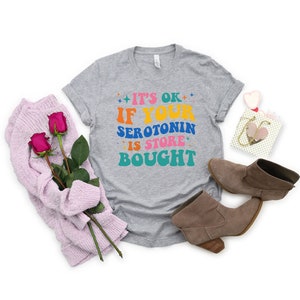 Puede incluir: Camiseta gris con el texto "It's ok if your serotonin is store bought" en letras de colores.