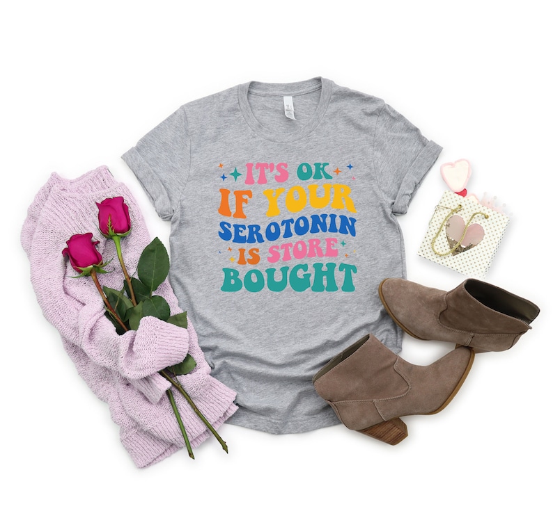 Puede incluir: Camiseta gris con el texto "It's ok if your serotonin is store bought" en letras de colores.