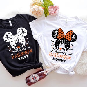 May include: A black t-shirt and a white t-shirt featuring a Mickey Mouse silhouette graphic with a castle, bats, and a witch on a broomstick. The text reads 2026 Disney HALLOWEEN DADDY on the black shirt and 2026 Disney HALLOWEEN MOMMY on the white shirt.