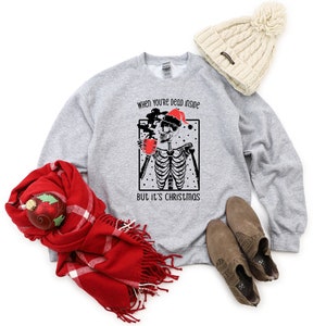 May include: A gray sweatshirt with a black and white illustration of a skeleton wearing a Santa hat and holding a mug. The text "When you're dead inside but it's Christmas" is printed on the sweatshirt.