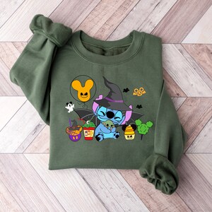 May include: Green sweatshirt with a blue Stitch character wearing a witch hat and holding a small green alien. The design includes a Mickey Mouse balloon, a ghost, and other Halloween-themed elements.