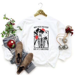 May include: White sweatshirt with a black and white illustration of a skeleton wearing a Santa hat and holding a cup of coffee. The text "When you're dead inside but it's Christmas" is printed on the sweatshirt.