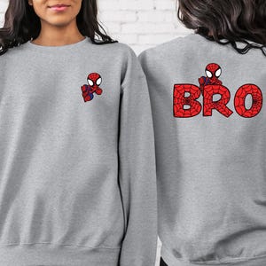 May include: Two grey sweatshirts with a Spiderman design. One sweatshirt has a small Spiderman graphic on the left chest and the other sweatshirt has a larger Spiderman graphic with the word "BRO" on the back.