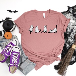 May include: A mauve t-shirt featuring a graphic of four cats dressed as ghosts. The cats are in a line, with the first two standing upright and the last two lying down. The t-shirt is on a white background with a pair of purple Converse shoes, a broom, and some Halloween decorations.