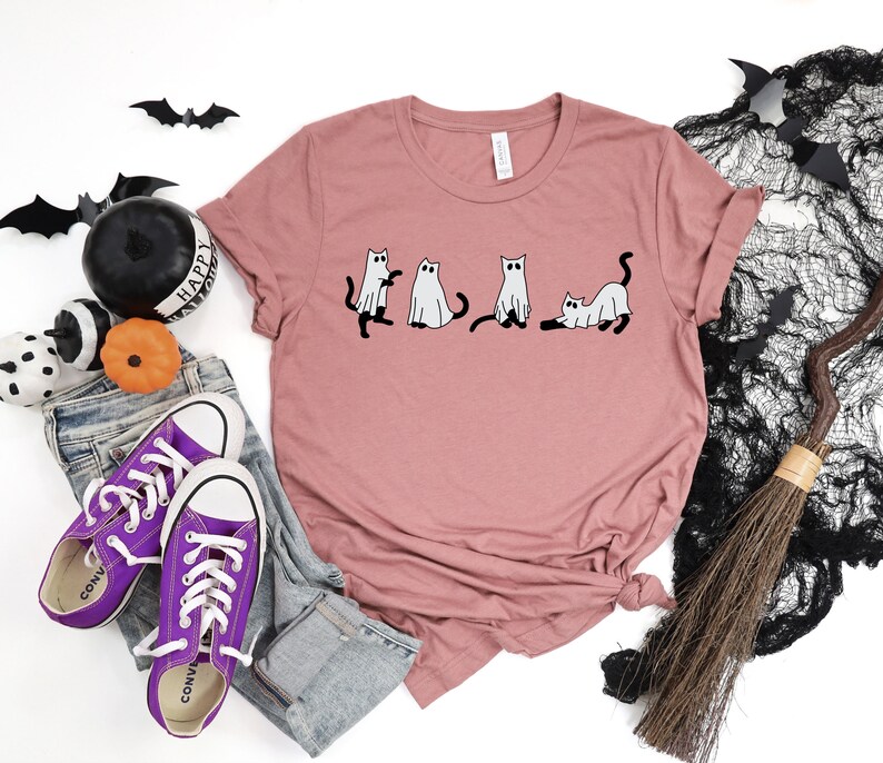 May include: A mauve t-shirt featuring a graphic of four cats dressed as ghosts. The cats are in a line, with the first two standing upright and the last two lying down. The t-shirt is on a white background with a pair of purple Converse shoes, a broom, and some Halloween decorations.