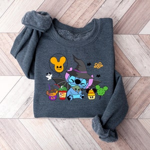 May include: A dark gray sweatshirt with a blue Stitch character wearing a witch hat and holding a small green monster. The design includes a Mickey Mouse head balloon, a witch's cauldron, and other Halloween-themed elements.