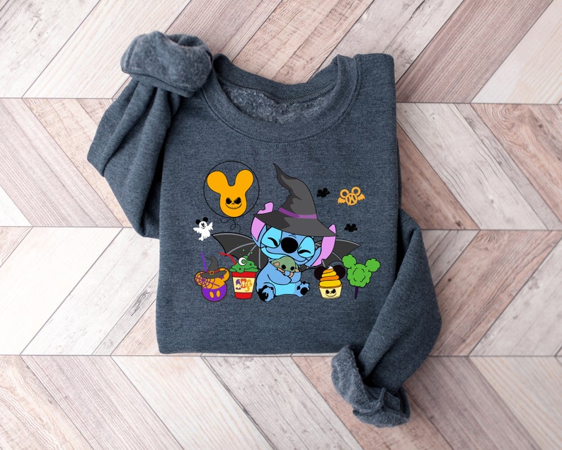 May include: A dark gray sweatshirt with a blue Stitch character wearing a witch hat and holding a small green monster. The design includes a Mickey Mouse head balloon, a witch's cauldron, and other Halloween-themed elements.