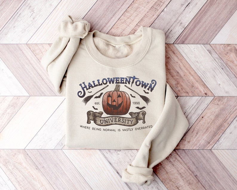 May include: A beige sweatshirt with a Halloween-themed graphic. The graphic features a pumpkin with a spooky face, bats, a broom, and the text "Halloweentown University est. 1998 Where being normal is vastly overrated".