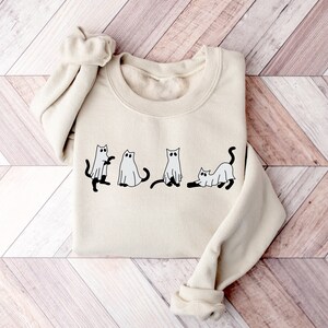 May include: A cream-coloured sweatshirt featuring a graphic of four cats dressed as ghosts. The cats are black with white ghost costumes and have black eyes.