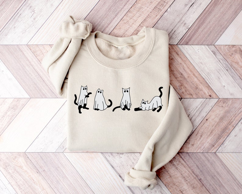 May include: A cream-coloured sweatshirt featuring a graphic of four cats dressed as ghosts. The cats are black with white ghost costumes and have black eyes.