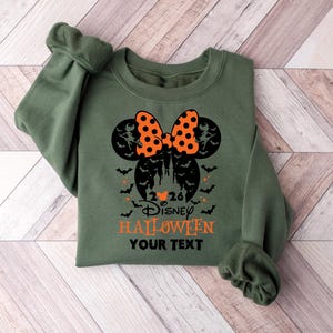 May include: An olive green crewneck sweatshirt featuring a black graphic of a castle silhouette inside a Mickey Mouse head shape, topped with an orange and black polka dot bow. The text reads 2026 Disney HALLOWEEN YOUR TEXT with bats.