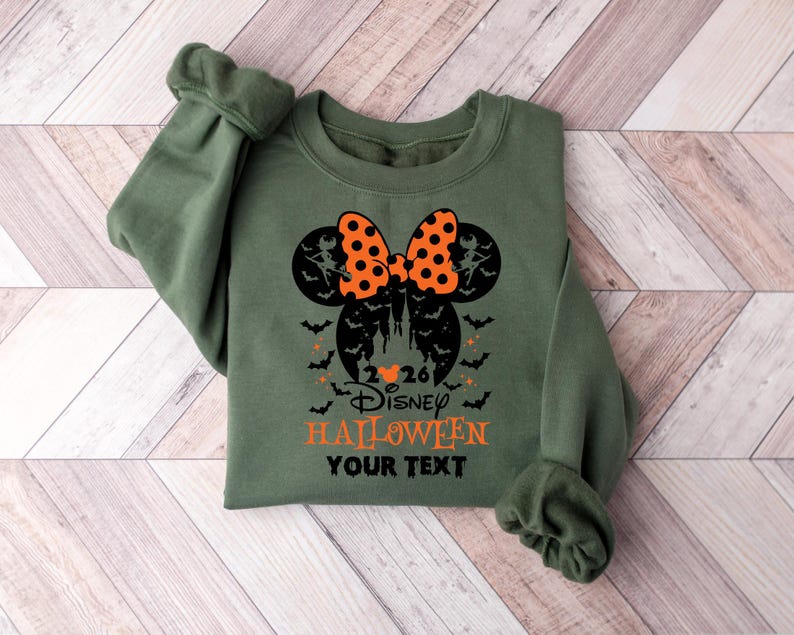 May include: An olive green crewneck sweatshirt featuring a black graphic of a castle silhouette inside a Mickey Mouse head shape, topped with an orange and black polka dot bow. The text reads 2026 Disney HALLOWEEN YOUR TEXT with bats.