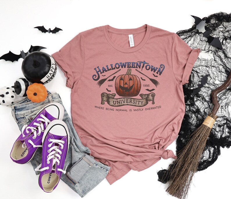 May include: A pink t-shirt with a graphic design of a pumpkin with a spooky face. The text on the shirt reads "Halloweentown University est. 1996" and "Where being normal is vastly overrated".