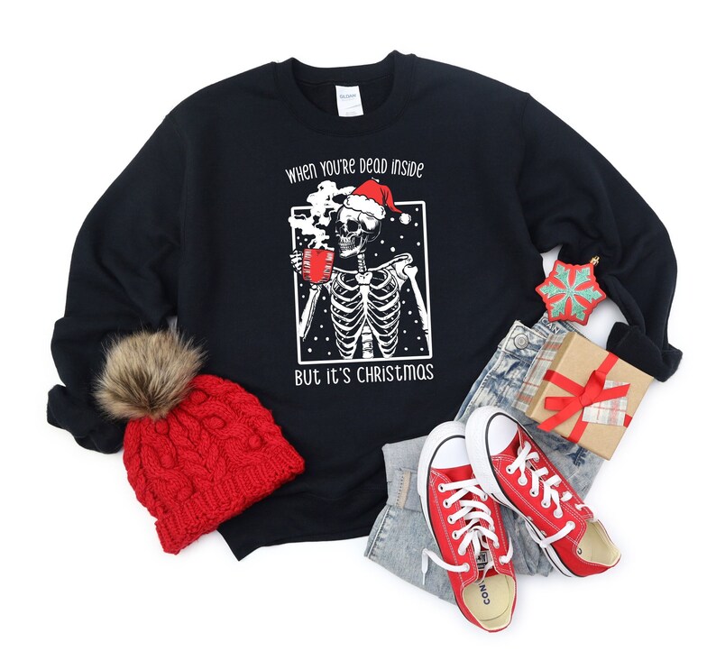 May include: Black Christmas sweater with a skeleton wearing a Santa hat and holding a mug. The text on the sweater reads "When you're dead inside but it's Christmas."