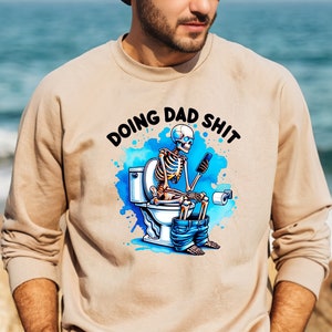 May include: Beige sweatshirt with the phrase "DOING DAD SHIT" above a cartoon skeleton on a toilet, using a phone. The design includes blue watercolor splashes and is a humorous take on fatherhood.