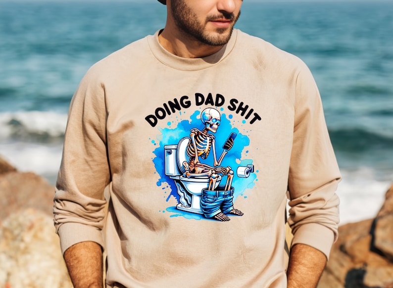 May include: Beige sweatshirt with the phrase "DOING DAD SHIT" above a cartoon skeleton on a toilet, using a phone. The design includes blue watercolor splashes and is a humorous take on fatherhood.