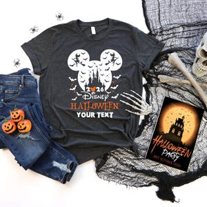 May include: A dark grey t-shirt featuring a white Mickey Mouse head silhouette containing a castle, bats, and a witch on a broomstick. The text reads 2026 Disney HALLOWEEN YOUR TEXT. Accompanied by blue jeans, three small pumpkins, and a skull.