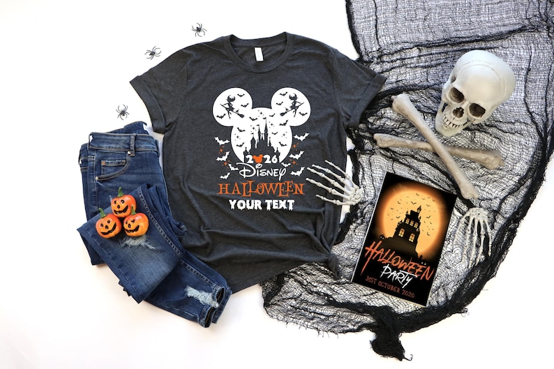 May include: A dark grey t-shirt featuring a white Mickey Mouse head silhouette containing a castle, bats, and a witch on a broomstick. The text reads 2026 Disney HALLOWEEN YOUR TEXT. Accompanied by blue jeans, three small pumpkins, and a skull.