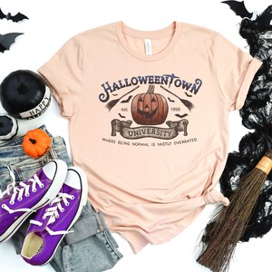 May include: A peach-colored t-shirt with a graphic design of a pumpkin with a face and the text "Halloweentown University est. 1996 Where being normal is vastly overrated".