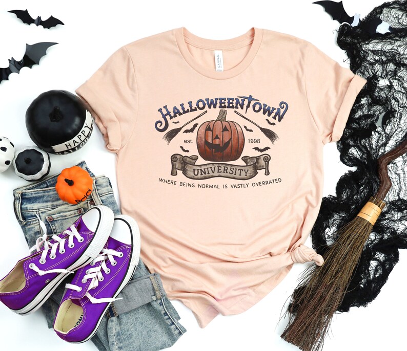 May include: A peach-colored t-shirt with a graphic design of a pumpkin with a face and the text "Halloweentown University est. 1996 Where being normal is vastly overrated".