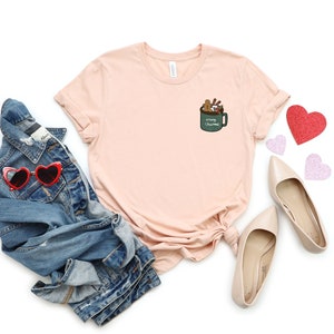 May include: A light pink t-shirt with a green mug graphic on the left chest. The mug has the text "Merry Christmas" and is filled with gingerbread cookies, candy canes, and marshmallows.