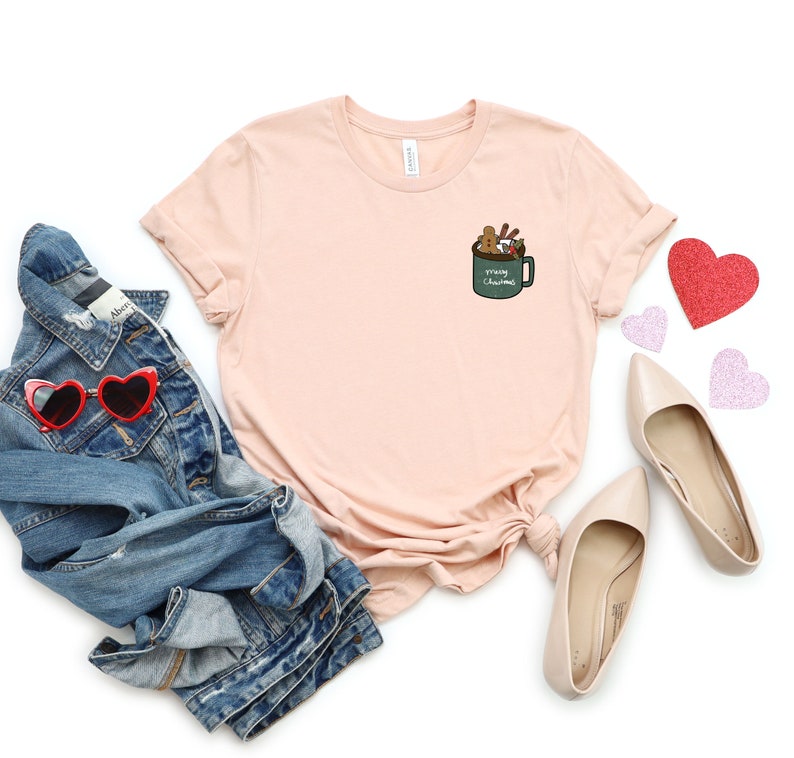 May include: A light pink t-shirt with a green mug graphic on the left chest. The mug has the text "Merry Christmas" and is filled with gingerbread cookies, candy canes, and marshmallows.
