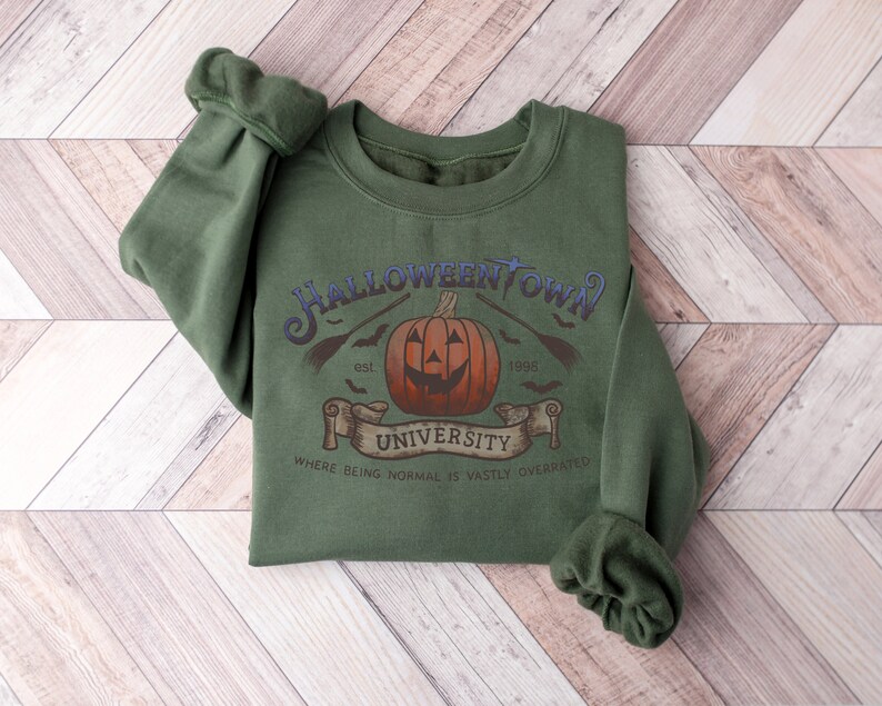 May include: A green sweatshirt with a Halloween-themed graphic. The graphic features a pumpkin with a jack-o'-lantern face, bats, and the text "Halloweentown University est. 1998. Where being normal is vastly overrated."