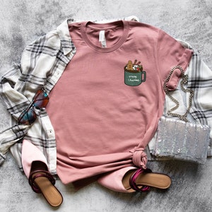 May include: A pink t-shirt with a graphic of a green mug with marshmallows, cinnamon sticks, and the text "'Tis the Season" on it. The shirt is laid flat on a gray background with a plaid shirt, sunglasses, a silver purse, and pink shoes.