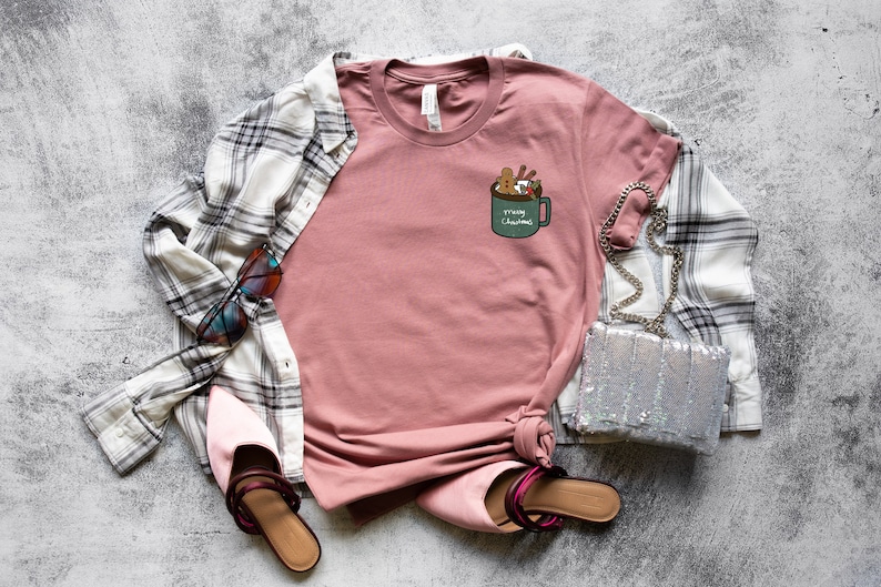 May include: A pink t-shirt with a graphic of a green mug with marshmallows, cinnamon sticks, and the text "'Tis the Season" on it. The shirt is laid flat on a gray background with a plaid shirt, sunglasses, a silver purse, and pink shoes.
