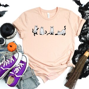 May include: A peach-coloured t-shirt featuring a graphic of four cats dressed as ghosts. The cats are in a line, with the first cat looking to the left, the second cat looking straight ahead, the third cat looking to the right, and the fourth cat lying down. The t-shirt is on a white background with a pair of purple Converse shoes, a pair of jeans, a black and white pumpkin, a black and white witch's hat, and a black and white broom.