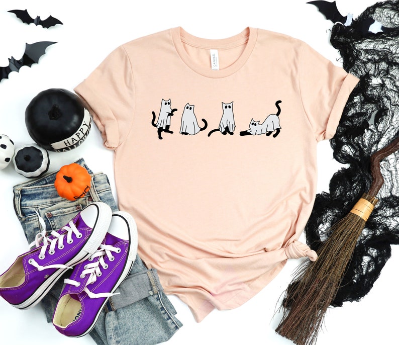 May include: A peach-coloured t-shirt featuring a graphic of four cats dressed as ghosts. The cats are in a line, with the first cat looking to the left, the second cat looking straight ahead, the third cat looking to the right, and the fourth cat lying down. The t-shirt is on a white background with a pair of purple Converse shoes, a pair of jeans, a black and white pumpkin, a black and white witch's hat, and a black and white broom.