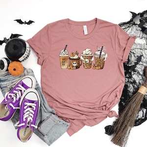 Pu&ograve; includere: Una maglietta color malva con una grafica di quattro bevande al caff&egrave; a tema Halloween. Le bevande sono decorate con zucche, fantasmi e pipistrelli. Il testo "Happy Halloween" &egrave; visibile sulla maglietta.