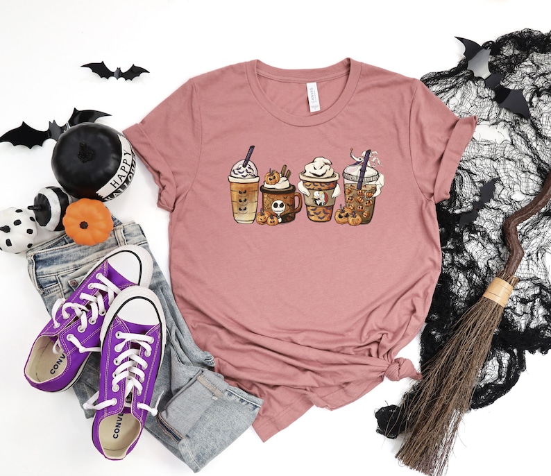 Pu&ograve; includere: Una maglietta color malva con una grafica di quattro bevande al caff&egrave; a tema Halloween. Le bevande sono decorate con zucche, fantasmi e pipistrelli. Il testo "Happy Halloween" &egrave; visibile sulla maglietta.
