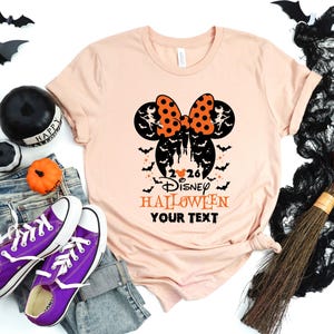 May include: A peach-coloured t-shirt featuring a black and orange graphic of mouse ears with a polka-dot bow, a castle silhouette, bats, and the text 2026 DISNEY HALLOWEEN YOUR TEXT. The design includes small witch figures and stars.