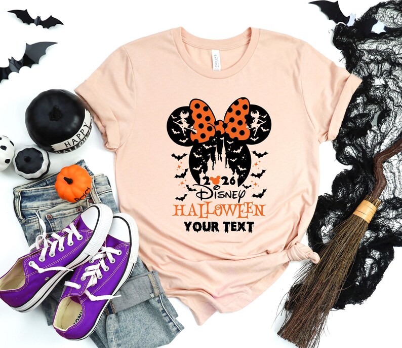 May include: A peach-coloured t-shirt featuring a black and orange graphic of mouse ears with a polka-dot bow, a castle silhouette, bats, and the text 2026 DISNEY HALLOWEEN YOUR TEXT. The design includes small witch figures and stars.