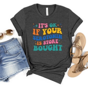 Puede incluir: Una camiseta gris oscuro con el texto "It's ok if your serotonin is store bought" en letras retro de colores.