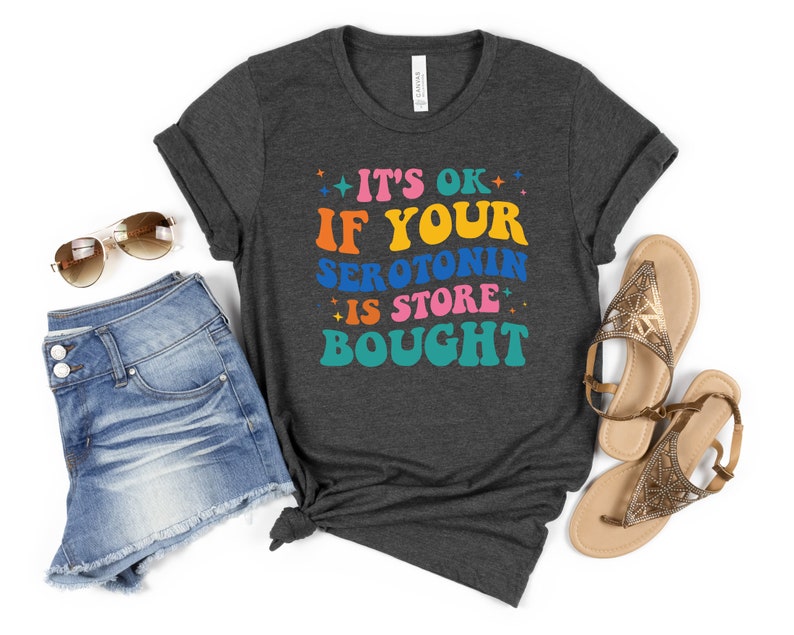 Puede incluir: Una camiseta gris oscuro con el texto "It's ok if your serotonin is store bought" en letras retro de colores.