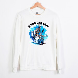 May include: White sweatshirt with the phrase "DOING DAD SHIT" above a colorful graphic of a skeleton on a toilet, using a phone. The graphic has blue watercolor-style accents.