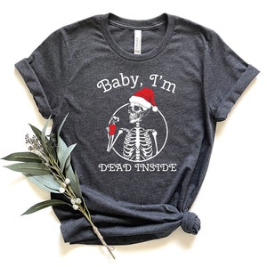 May include: A dark gray t-shirt with a white skeleton wearing a Santa hat and holding a drink. The text "Baby, I'm Dead Inside" is printed on the shirt.