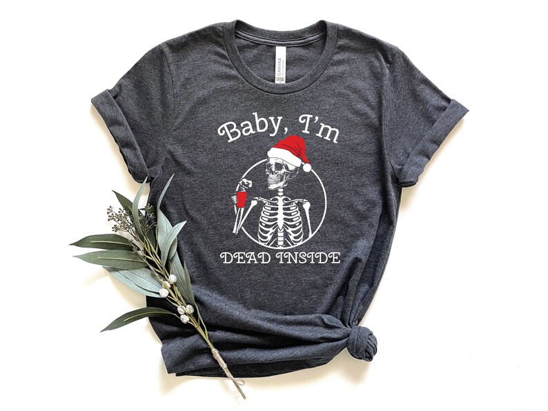 May include: A dark gray t-shirt with a white skeleton wearing a Santa hat and holding a drink. The text "Baby, I'm Dead Inside" is printed on the shirt.