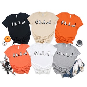 May include: Five t-shirts featuring a black and white graphic of a line of cats dressed as ghosts. The shirts are in various colours: black, beige, orange, white and grey.