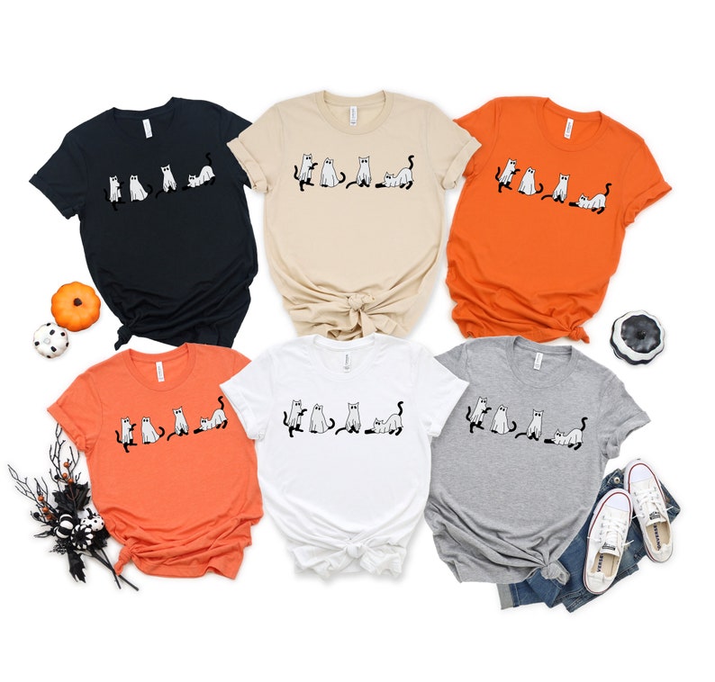 May include: Five t-shirts featuring a black and white graphic of a line of cats dressed as ghosts. The shirts are in various colours: black, beige, orange, white and grey.
