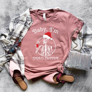 May include: A pink t-shirt with a white graphic of a skeleton wearing a Santa hat and holding a glass of red liquid. The text "Baby, I'm Dead Inside" is printed below the skeleton.