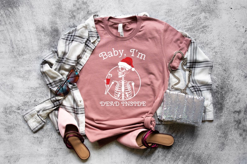 May include: A pink t-shirt with a white graphic of a skeleton wearing a Santa hat and holding a glass of red liquid. The text "Baby, I'm Dead Inside" is printed below the skeleton.