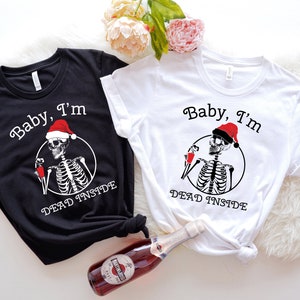 May include: Two black and white t-shirts with a skeleton wearing a Santa hat and holding a glass of wine. The text on the shirts reads "Baby, I'm Dead Inside".