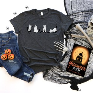 May include: A dark heather grey t-shirt featuring a graphic of five black cats dressed as ghosts. The shirt is laid out on a white surface with a pair of blue jeans, three orange jack-o'-lantern pumpkins, and Halloween decorations.