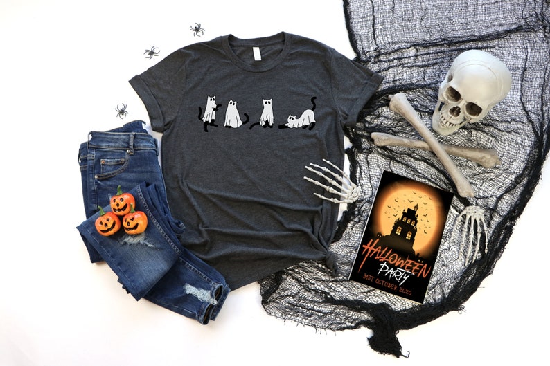May include: A dark heather grey t-shirt featuring a graphic of five black cats dressed as ghosts. The shirt is laid out on a white surface with a pair of blue jeans, three orange jack-o'-lantern pumpkins, and Halloween decorations.