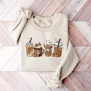 Pu&ograve; includere: Una felpa beige con una grafica di quattro bevande al caff&egrave; a tema Halloween. Le bevande sono decorate con zucche, fantasmi e altre immagini di Halloween.