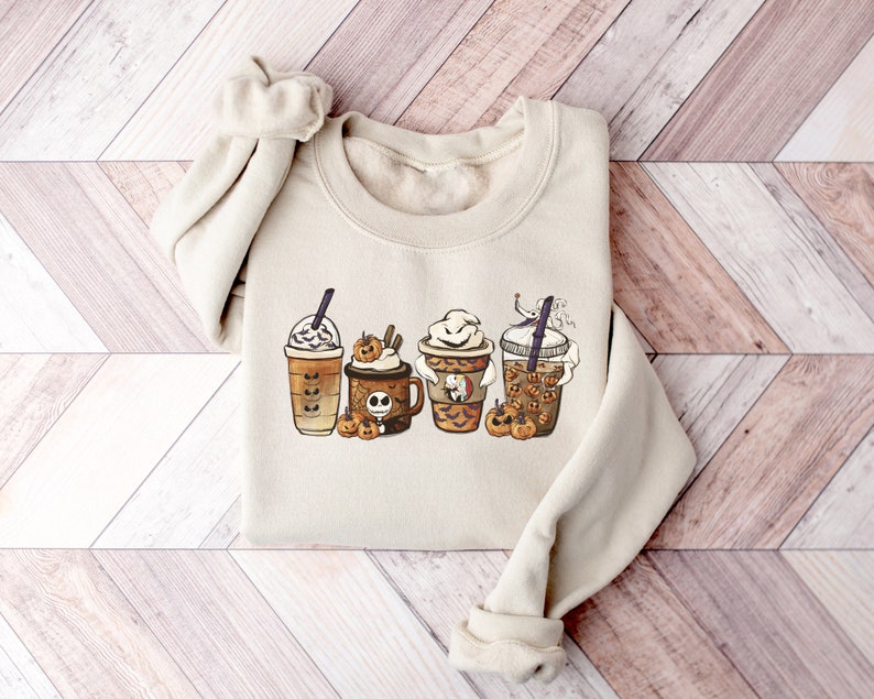 Pu&ograve; includere: Una felpa beige con una grafica di quattro bevande al caff&egrave; a tema Halloween. Le bevande sono decorate con zucche, fantasmi e altre immagini di Halloween.