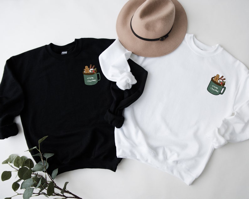 May include: Two sweatshirts, one black and one white, with a graphic of a green mug filled with Christmas treats and the text "Merry Christmas".
